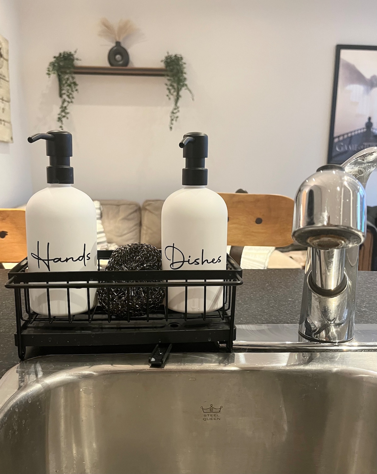 A kitchen sink area is displayed, featuring a black wire caddy that holds two labeled soap dispensers for hands and dishes. A clean stainless steel faucet is visible, alongside a sponge. The background shows a soft seating area, contributing to a functional kitchen space.