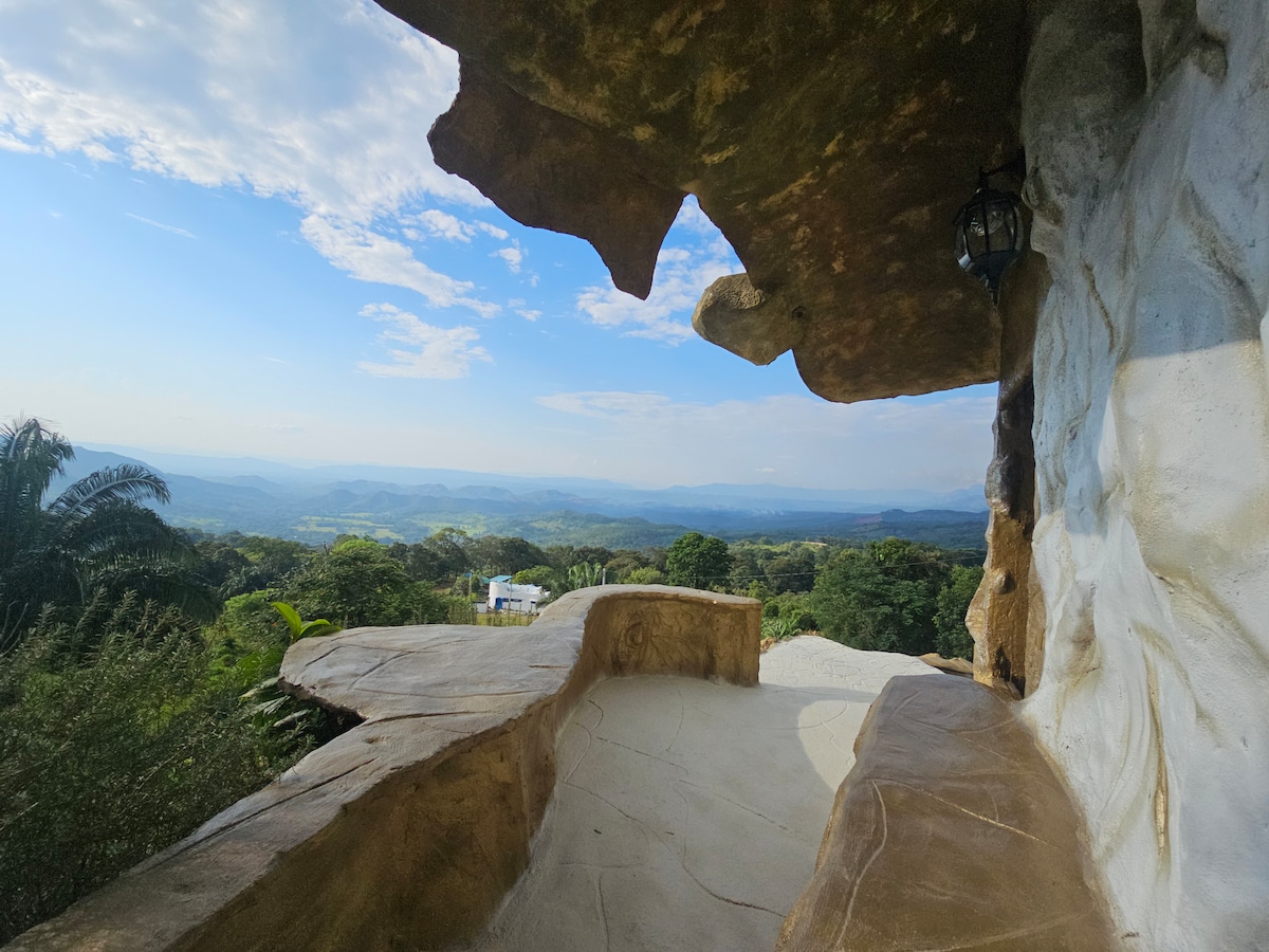 An expansive outdoor terrace is set against a backdrop of rolling hills and a clear sky. Unique rock formations frame the space, which features a smooth, curved seating area. Lush greenery can be seen in the distance, enhancing the natural surroundings.
