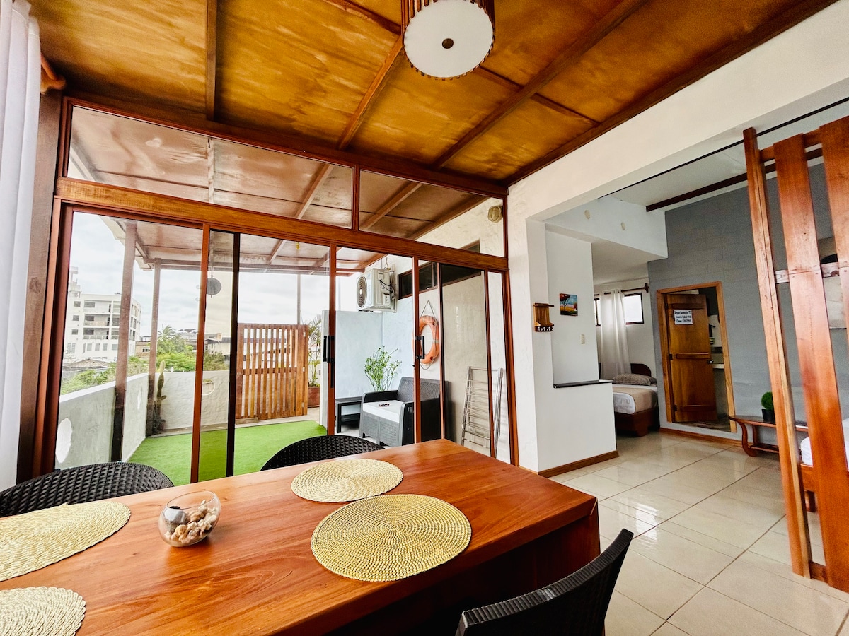 A dining area features a wooden table surrounded by black wicker chairs, with a natural fiber placemat on the table. Large glass sliding doors open to a terrace space, revealing greenery and natural light throughout the room.