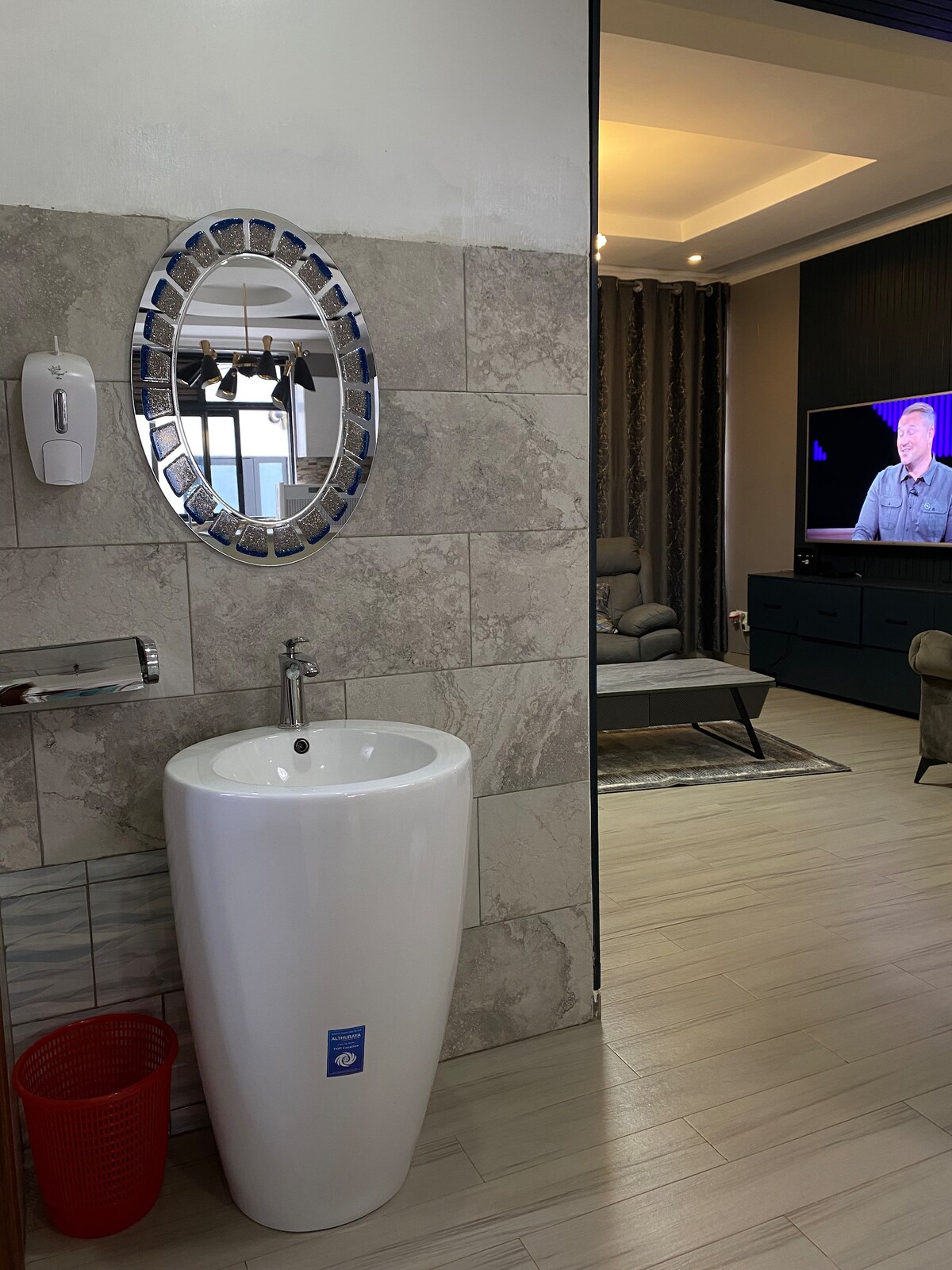 A modern bathroom area features a sleek, white pedestal sink below a round mirror with decorative framing. A soap dispenser is mounted on the wall nearby, while a red waste bin and a glimpse of a cozy living area with a television can be seen in the background.