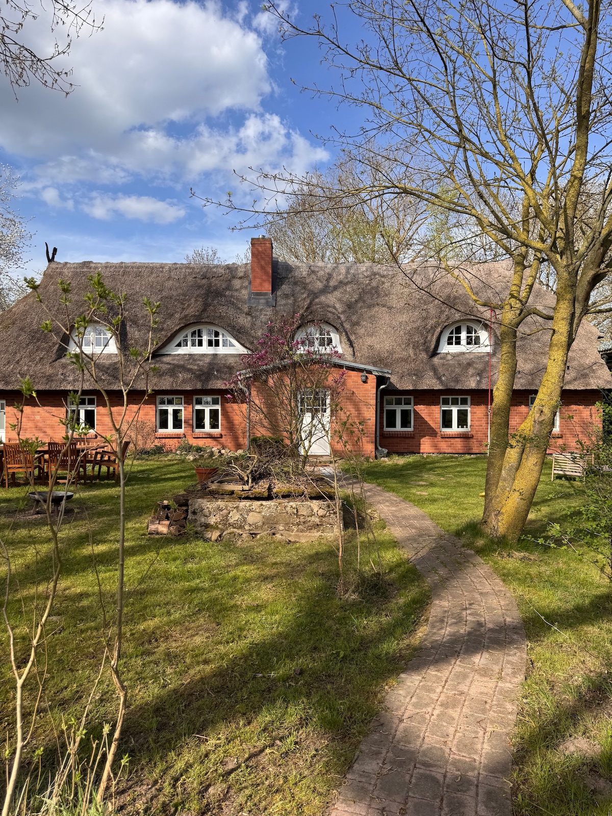The house features a thatched roof and red brick exterior, surrounded by a green lawn and trees. A stone pathway leads to the entrance, with benches and a dining set placed outdoors, offering a serene view of the natural surroundings.