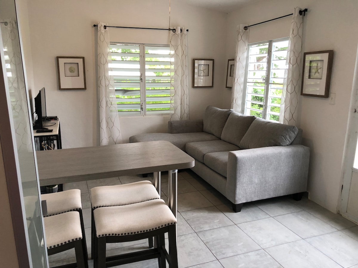 A light-filled living area features a comfortable gray sofa and a dining table with stools. Large windows with horizontal slats allow for natural light, while framed artwork decorates the white walls, enhancing the serene ambience.