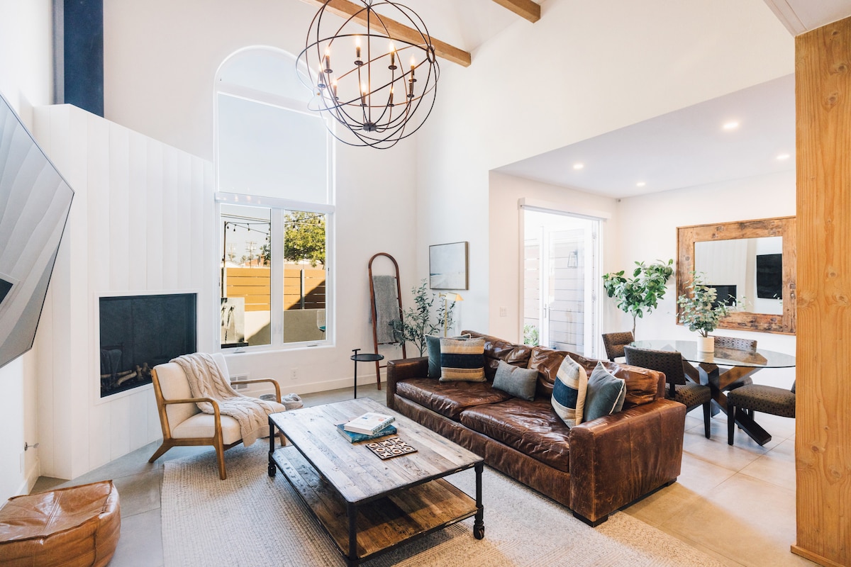 A spacious living area features high ceilings and large windows allowing natural light to fill the room. A leather sectional and stylish accent chair surround a wooden coffee table, while a dining table is positioned nearby. Decorative plants and modern lighting add to the atmosphere.