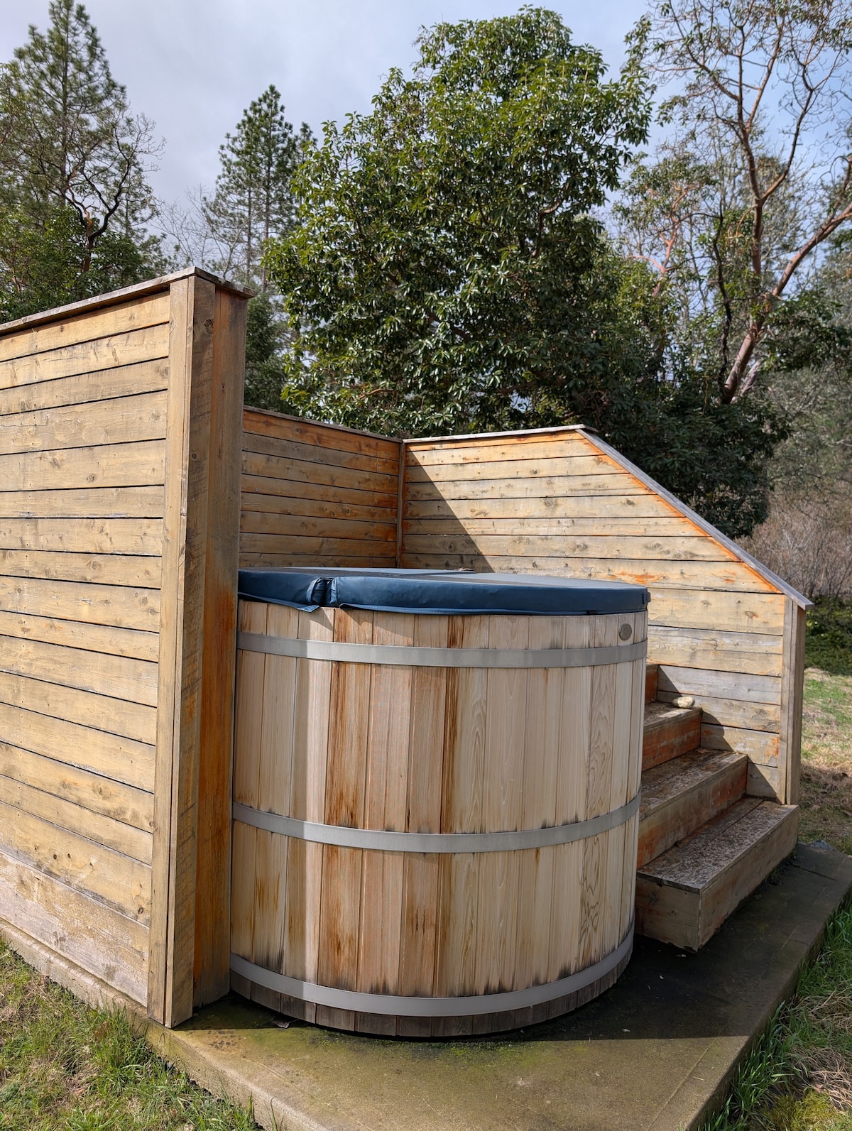 An outdoor wooden hot tub is nestled in a wooden enclosure, featuring steps leading up to it. The tub is round and has a blue cover. Surrounding greenery adds a natural touch to the serene setting.
