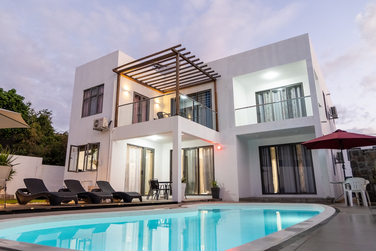 A modern villa is showcased, featuring a spacious patio area adjacent to a pristine pool. Comfortable loungers are positioned near the water, and tall glass doors provide a view into the inviting interior. An umbrella offers shade over an outdoor seating area.