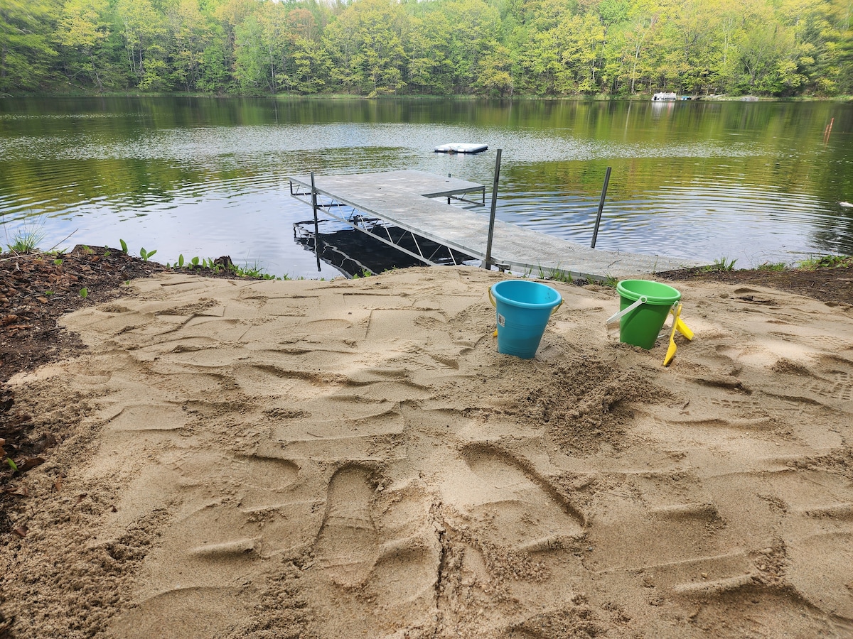 The image shows a sandy beach area leading to a tranquil lake. Two colorful buckets are placed in the sand, alongside a yellow shovel. A private pier extends over the water, surrounded by lush greenery.