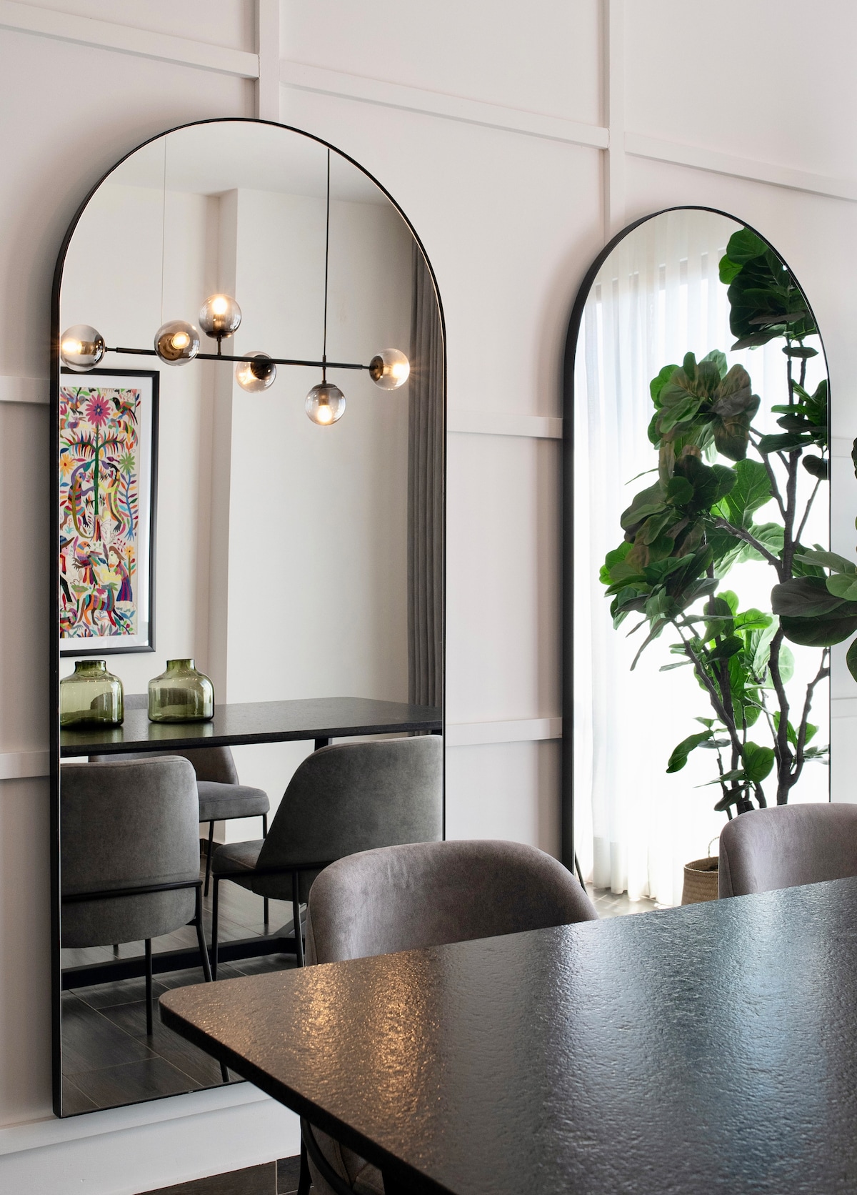 A modern dining area is featured with two arched mirrors reflecting stylish lighting fixtures and a nearby potted plant. A sleek table is surrounded by soft, grey chairs, creating an inviting space enhanced by natural light filtering through sheer curtains.
