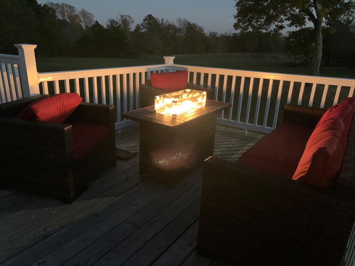 A spacious deck features a fire table surrounded by two cozy seating areas, each adorned with red cushions. Soft glow light illuminates the setting, creating a warm ambiance against a backdrop of rolling pastures and evening sky.