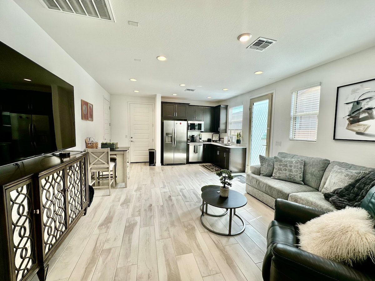 A spacious living area is featured, showcasing a comfortable sofa and a round coffee table. Dark cabinetry provides contrast to the light-colored flooring. The kitchenette is equipped with modern appliances, and natural light enters through large windows, creating a bright and airy environment.