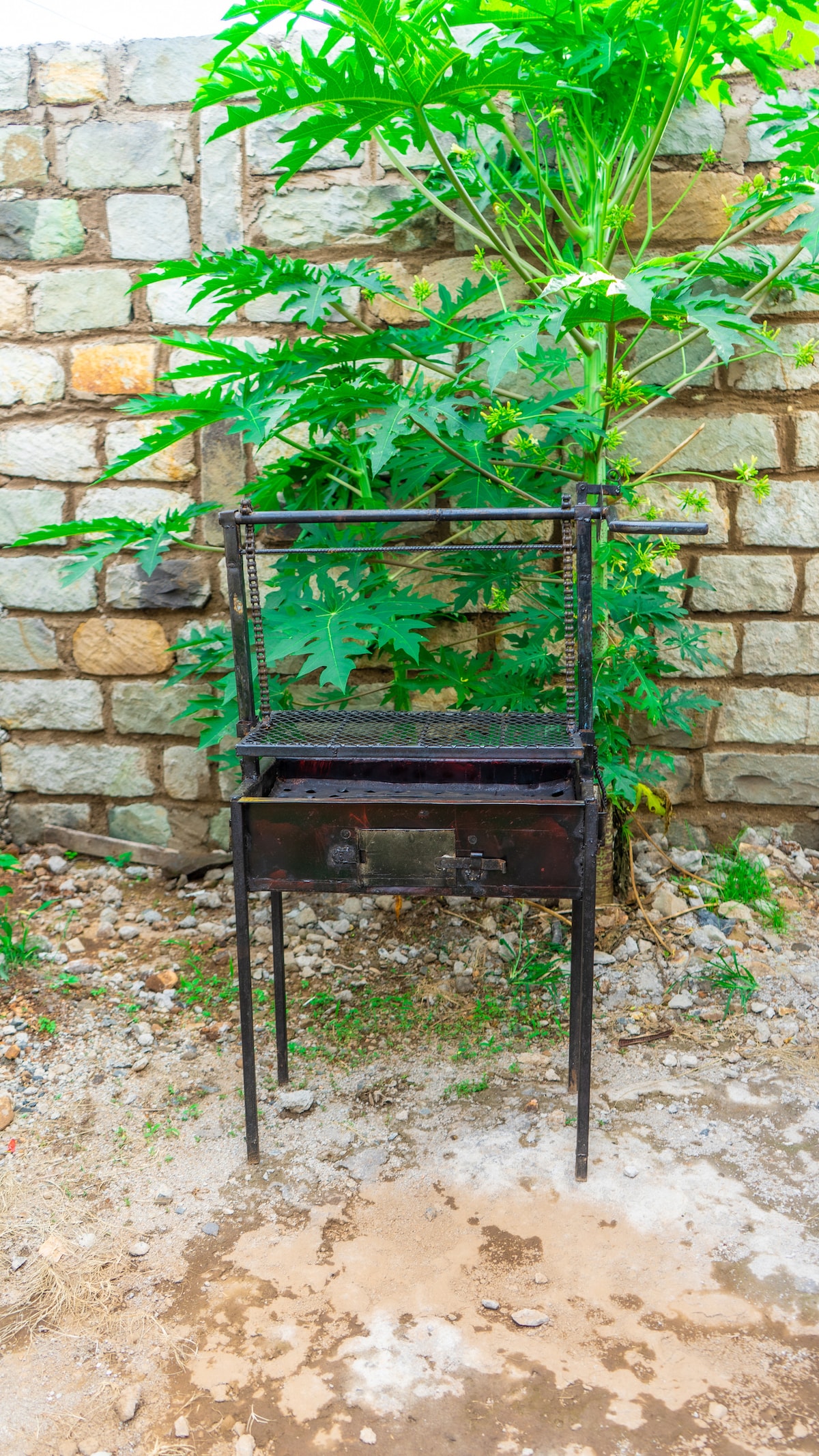 A metal barbecue grill stands in a garden setting next to a stone wall. Lush green foliage is visible in the background, providing a natural ambiance. The grill features a cooking surface elevated on sturdy legs, inviting opportunities for outdoor cooking and gatherings.