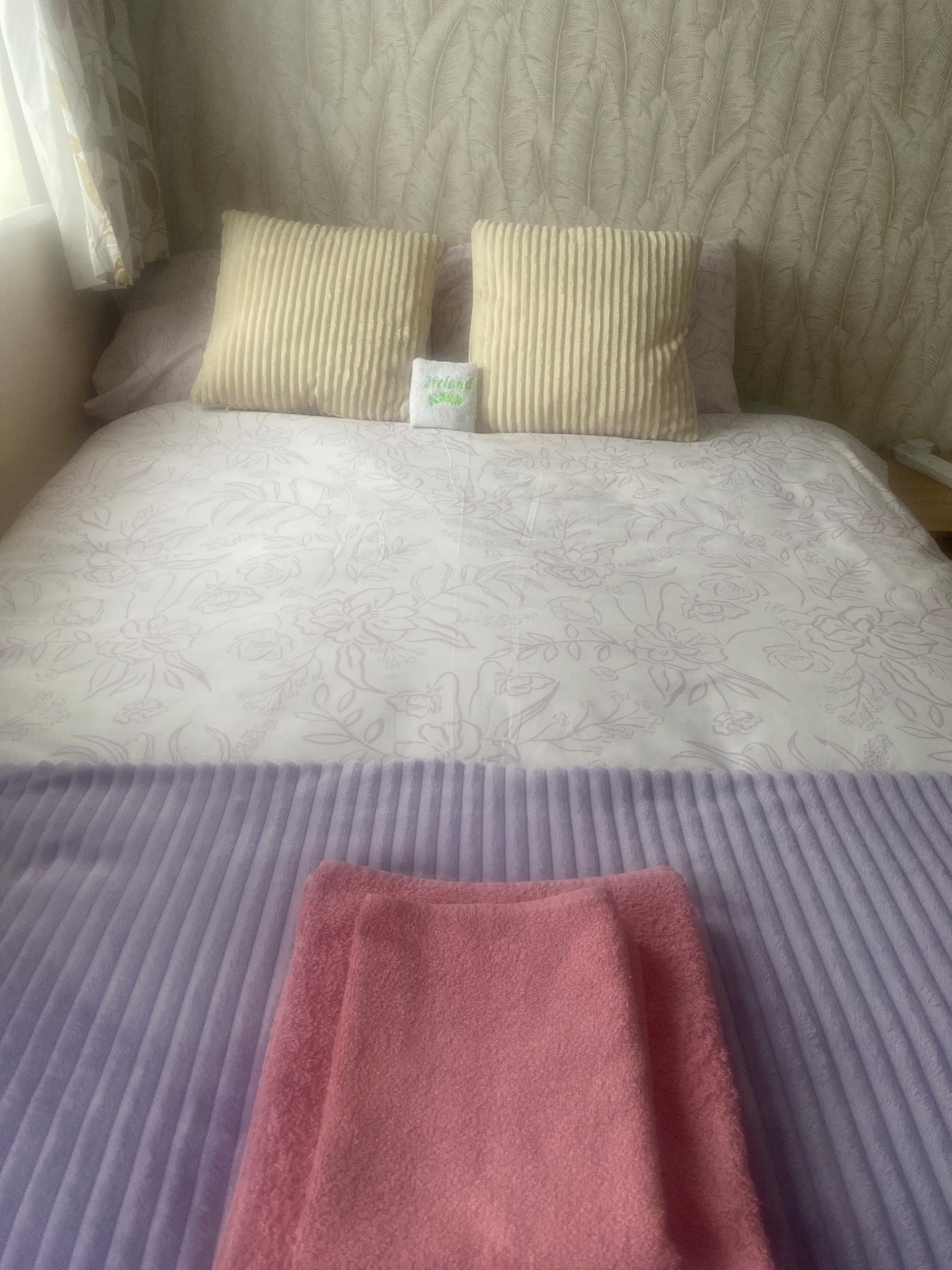 The sleeping area features a spacious bed dressed in floral-patterned bedding. Two striped cushions are positioned at the head of the bed. A folded pink towel is placed neatly at the foot of the bed, adding a touch of color to the arrangement.