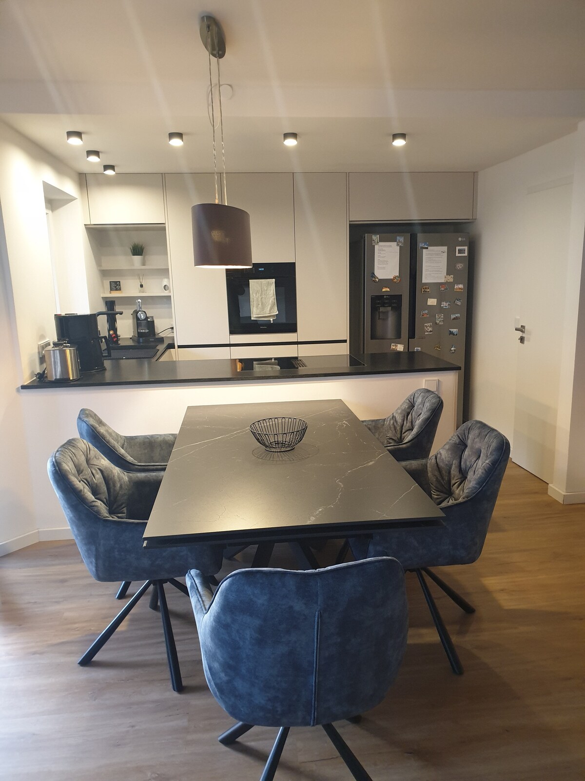 A modern kitchen and dining area are presented, featuring a sleek black table surrounded by four upholstered chairs. A stylish pendant light hangs above the table. Kitchen appliances, including a refrigerator and an oven, are visible in the background, enhancing the functional design of the space.