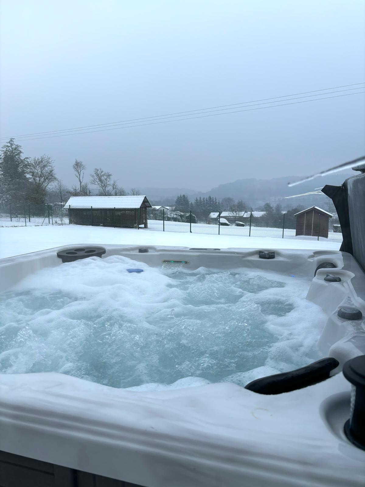 A hot tub is filled with bubbling water, surrounded by a snowy landscape. Distant wooden structures are visible, set against a backdrop of gently rolling hills. The sky appears overcast, contributing to a serene winter atmosphere.