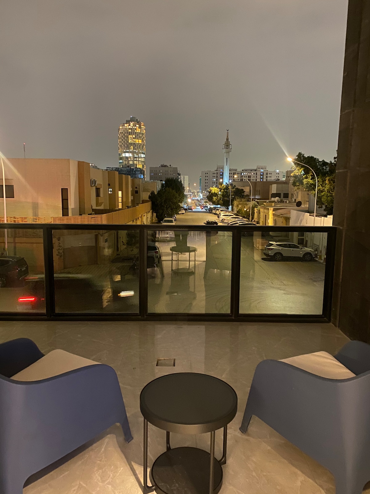 A private balcony is furnished with two modern blue chairs and a small round table. A city view is visible, featuring low-rise buildings and a distant tower, all illuminated against the night sky.