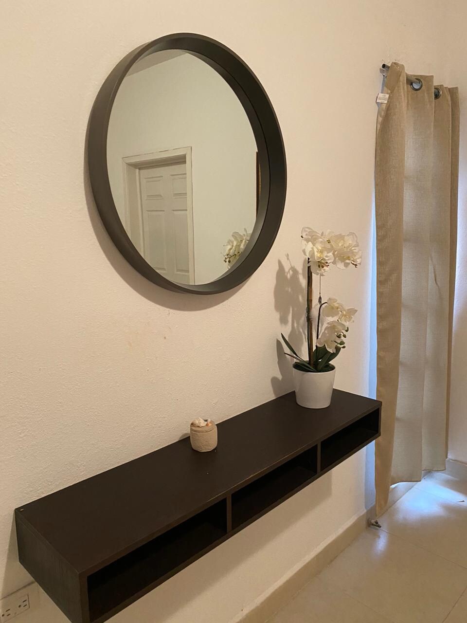 A round mirror is mounted on a white wall above a sleek, dark console table. A small potted orchid and a candle sit on the table, adding a touch of elegance. A light curtain partially frames a window, allowing natural light to enter the space.