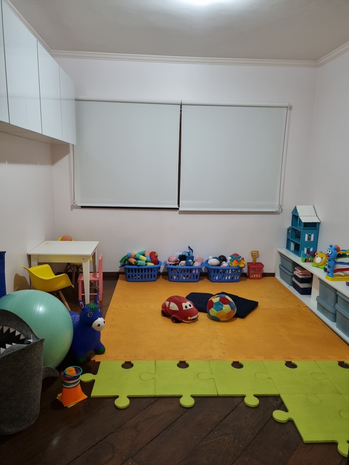 A colorful playroom is designed for children, featuring a bright orange carpet and various toys arranged in baskets along the wall. A small table and chairs are positioned beside a green puzzle mat, adding to the inviting atmosphere of the space.