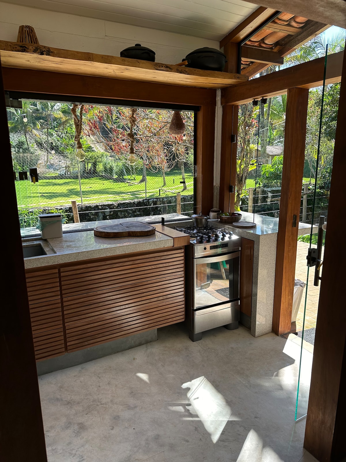 A well-equipped kitchen is highlighted by natural light streaming through large glass windows. Wooden cabinetry contrasts with modern appliances. The space offers a view of the lush greenery outside, creating an inviting atmosphere for meal preparation.