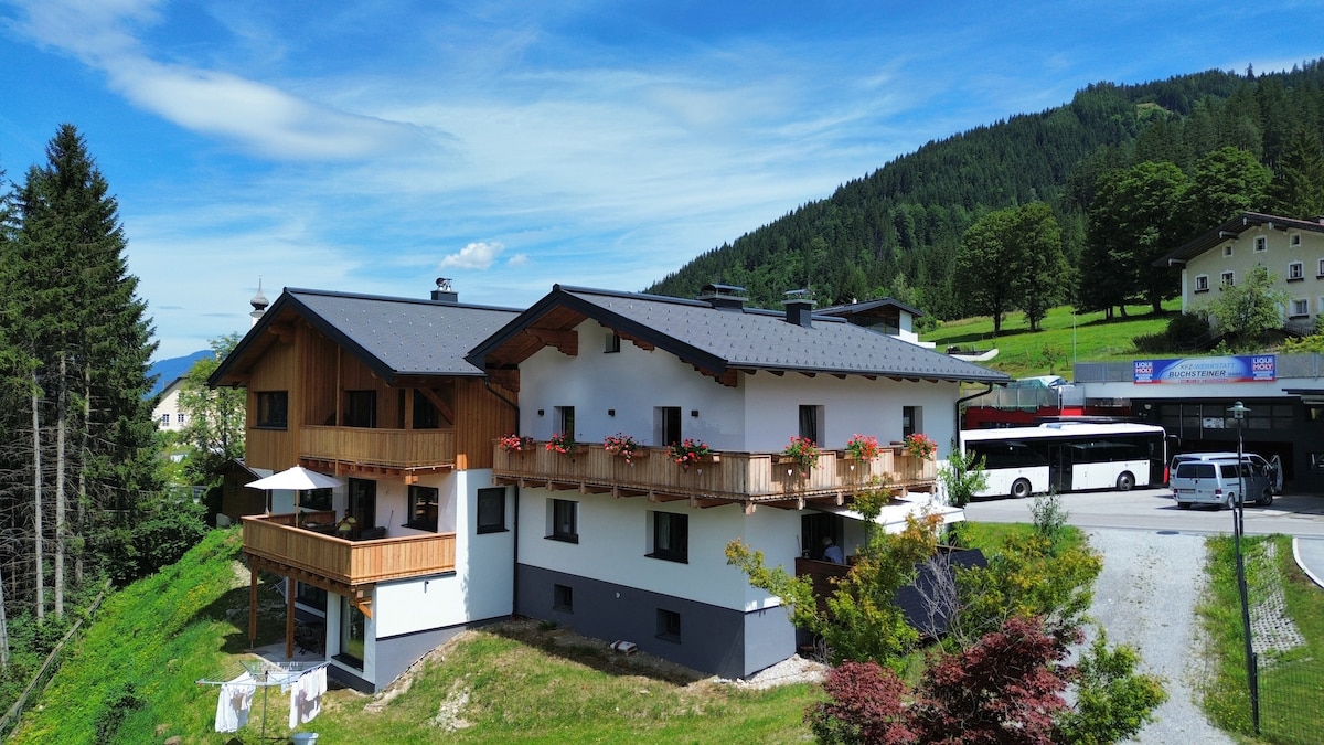 The chalet features a combination of wood and white exterior with balconies adorned with vibrant flower boxes. Surrounded by greenery, the building is situated in a serene mountain landscape, with a bus stop visible nearby for convenient access.