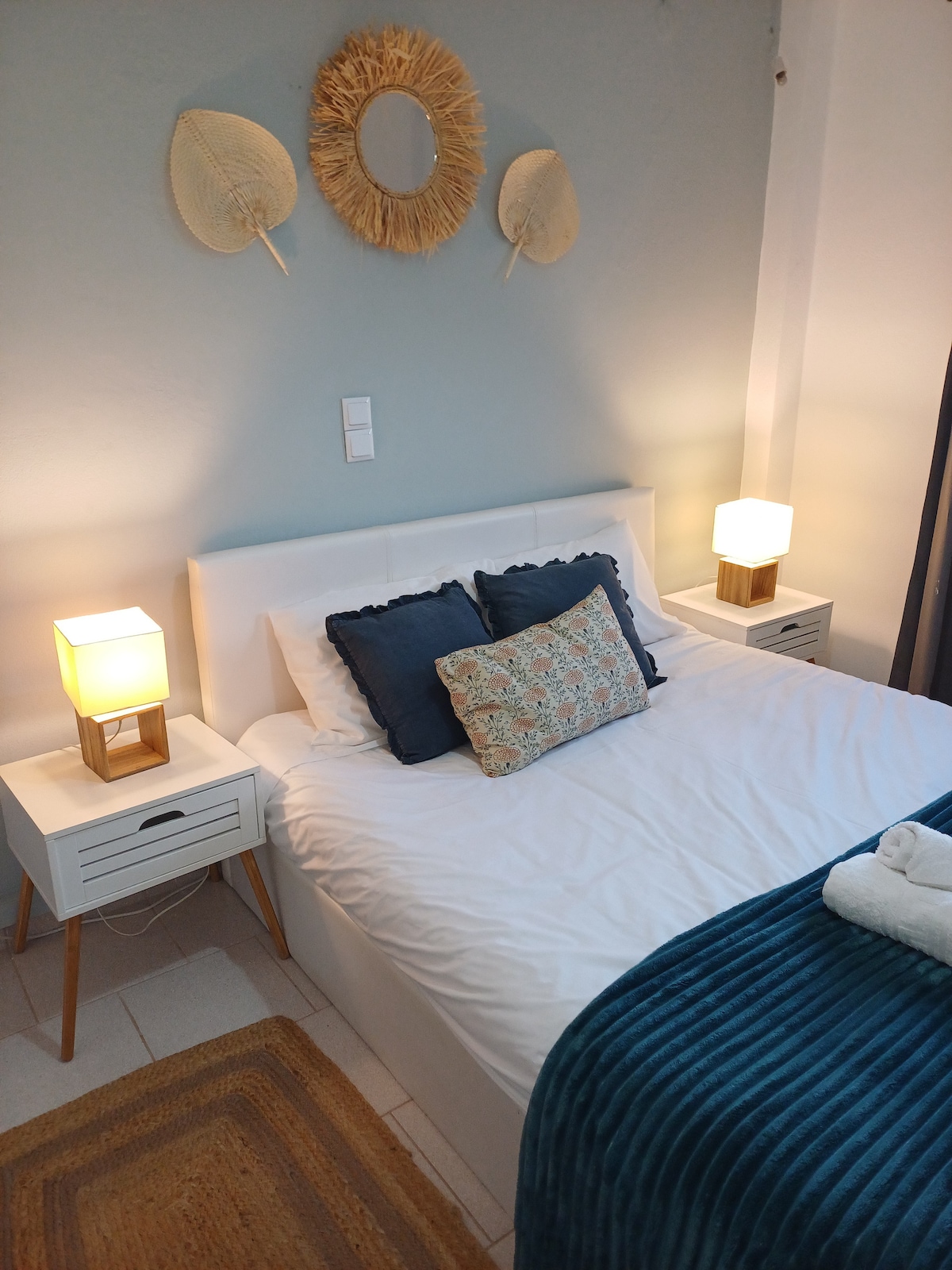 A welcoming sleeping area is presented with a spacious bed dressed in crisp white linens. Two bedside tables with lamps frame the bed, providing soft lighting. Decorative pillows add a touch of color, while a round mirror hangs above, complementing the soothing wall color.