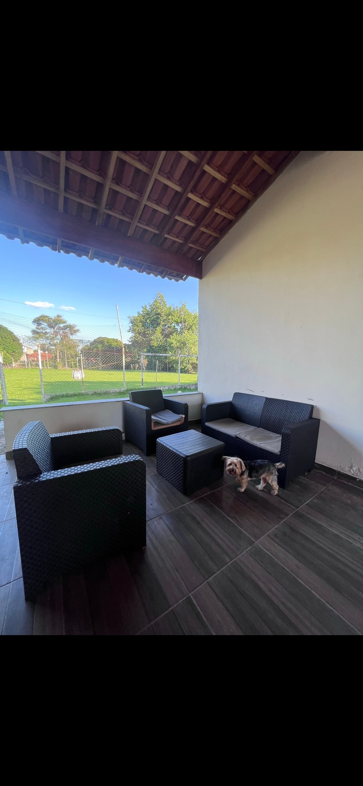 An outdoor seating area features modern furniture in dark tones, including a sofa, two armchairs, and a small table. Natural light filters through an open space, highlighting a grassy field visible in the background, with trees lining the edge.