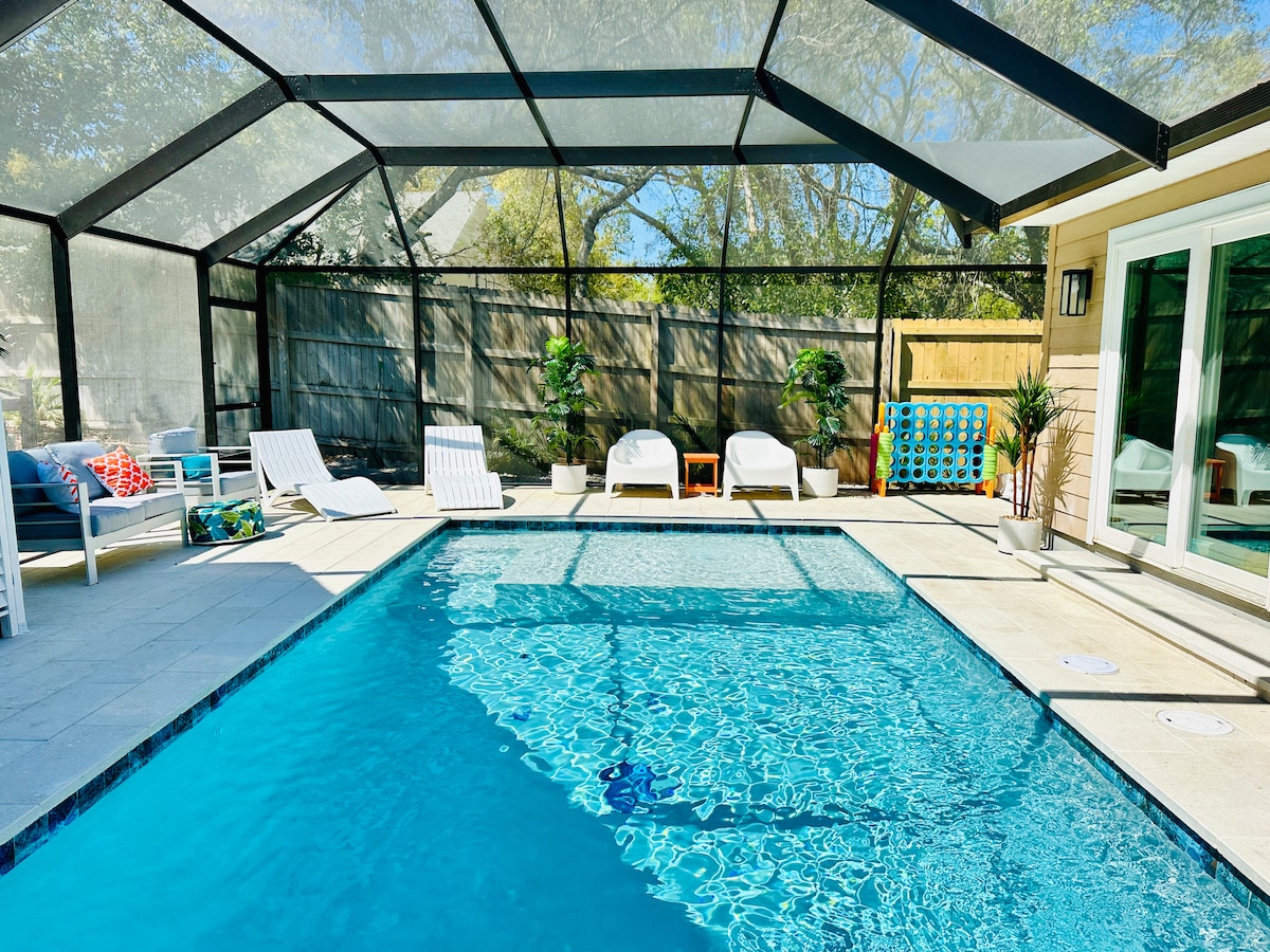A heated enclosed pool is featured, surrounded by a spacious patio area. Poolside loungers are positioned for relaxation, while dappled sunlight reflects off the clear water. A colorful game board adds a playful touch in the background, set against a backdrop of tall greenery.