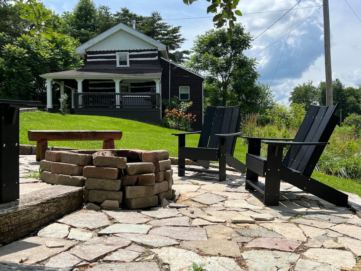 A stone patio surrounds a fire pit made of bricks, with two black Adirondack chairs positioned for comfort. In the background, the cabin's charming facade is visible, complemented by lush greenery and a cloudy sky.