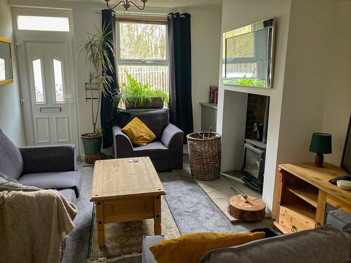 The open-plan living area features a cozy seating arrangement with a grey sofa and a dark armchair. A wooden coffee table sits in front, while a decorative plant in the corner adds a touch of greenery. A wood-burner stove is positioned against a stone wall, providing warmth.