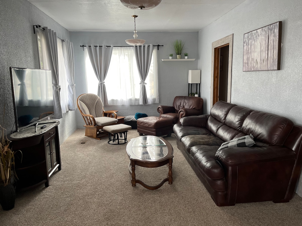 A well-appointed living room features a dark leather sectional sofa and an armchair, accompanied by a wooden coffee table. Natural light enters through large windows adorned with sheer curtains, adding to the room's brightness. A cozy accent chair is positioned nearby, enhancing the inviting space.