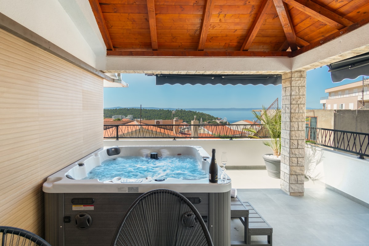 An outdoor terrace features a hot tub beneath a wooden pergola, showcasing panoramic views of the coastline. A bottle of champagne and two glasses are positioned on a small table nearby, while potted plants add a touch of greenery to the space.