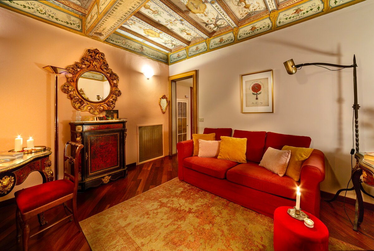 A richly decorated living room features a vibrant red sofa adorned with neutral cushions and is complemented by a round side table with a soft glow from a candle. Elegant hand-painted ceiling frescoes add historical charm above, while antique furniture reflects a classic style.