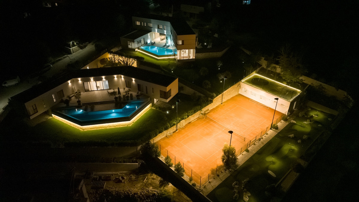 An aerial view captures the illuminated modern villas, each featuring swimming pools. A well-lit orange clay tennis court is highlighted adjacent to the expansive green landscape, which includes landscaped areas and pathways, creating a serene nighttime ambiance.