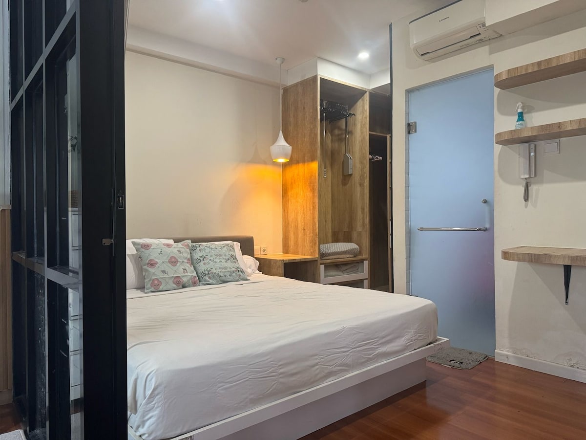 A spacious sleeping area features a king-size bed with decorative pillows against a light wall. A modern pendant light illuminates the room, while a minimalist closet and open shelving provide convenient storage options. A sleek door leads to the bathroom area.