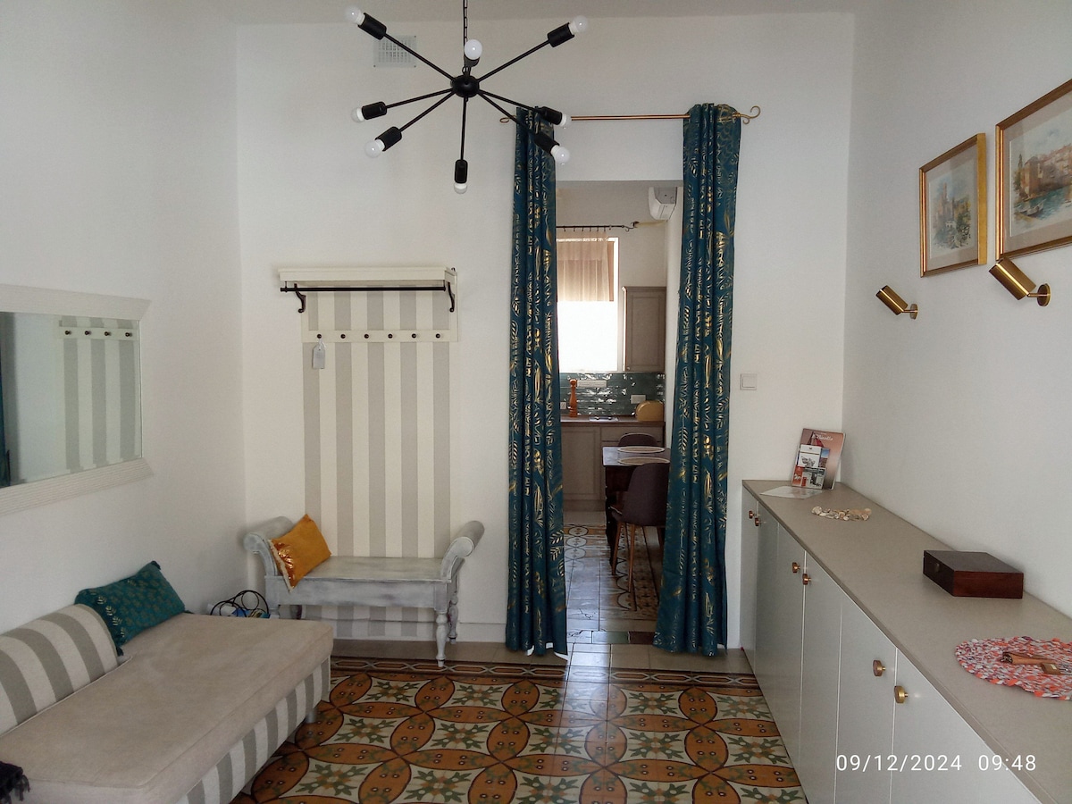 A section of the living space is displayed, featuring a light-colored sofa against a wall with vertical stripes. Colorful curtains frame a doorway leading to the kitchen area. Decorative tiles cover the floor, and a mirror and artwork are presented on adjacent walls.