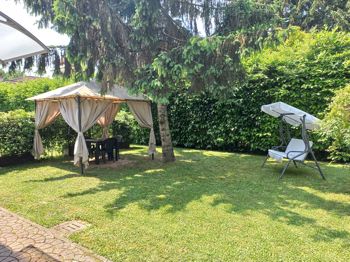 A spacious garden area features a gazebo with draped fabric, accompanied by a table and chairs. A lounging chair is positioned nearby, surrounded by lush greenery and a well-maintained lawn, offering a serene outdoor space for relaxation.