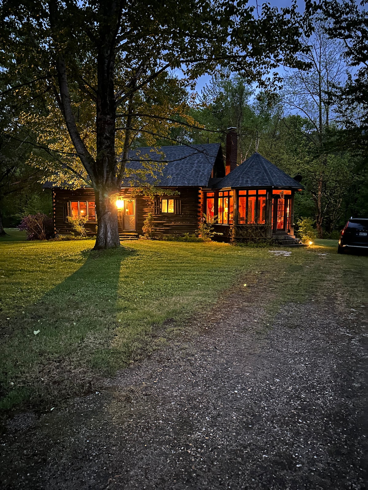 A charming log cabin is softly illuminated by warm lights as dusk settles. Surrounded by lush greenery, the cabin features large windows that highlight the cozy interior. A gravel driveway leads to the entrance, enhancing the tranquil setting.