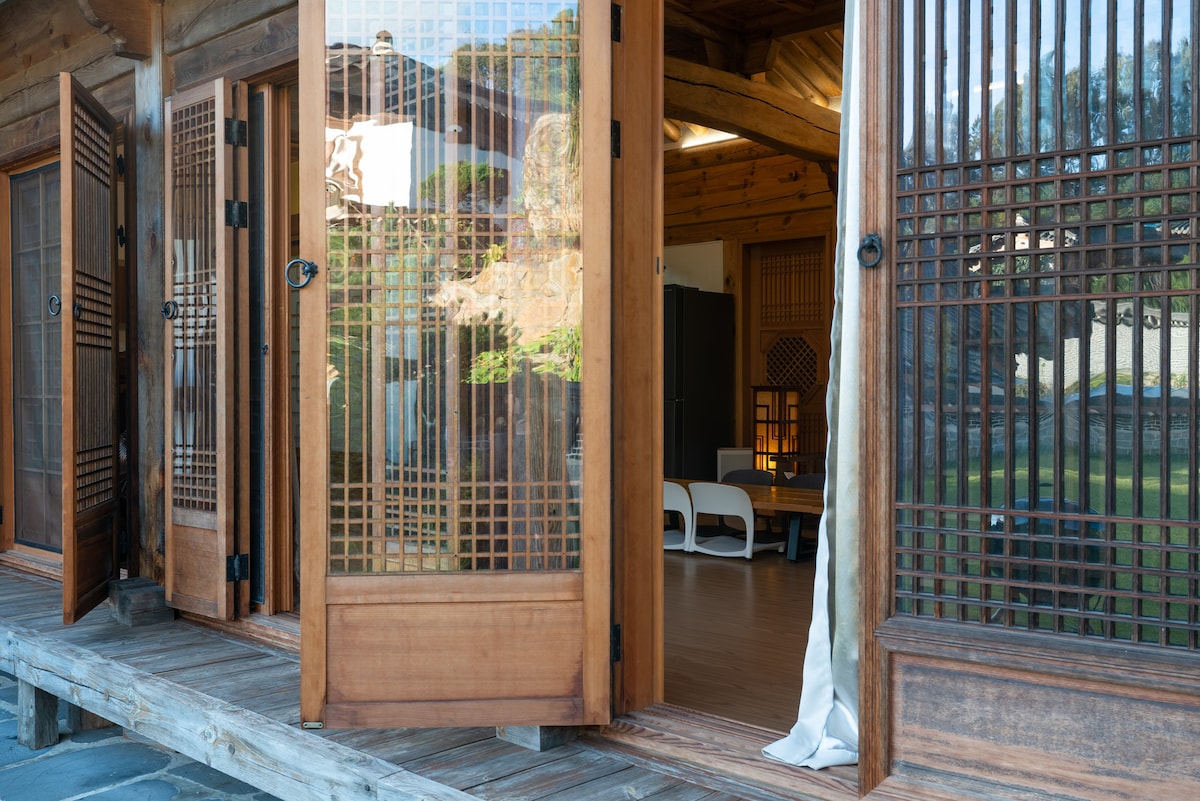 Traditional wooden doors with intricate lattice designs are open, revealing a spacious interior filled with natural light. The entrance leads to a wooden deck overlooking a lush green yard, inviting fresh air and a sense of tranquility.