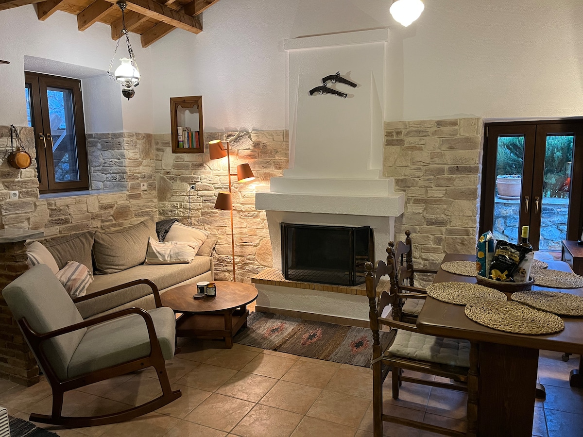 A cozy living area is highlighted by a stone fireplace and vintage wooden furniture. A comfortable sofa and cozy armchair invite relaxation. Natural light enters through windows, revealing a rustic charm that blends warmth with simplicity. A dining table is set for meals, completing this inviting space.