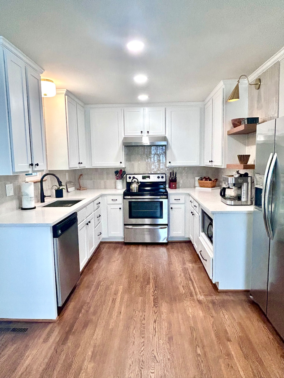 A newly remodeled kitchen showcases sleek white cabinetry and modern stainless steel appliances. Natural light illuminates the space, highlighting a spacious countertop that offers ample cooking area. Open shelves display decorative items, and a coffee maker is positioned conveniently on the counter.