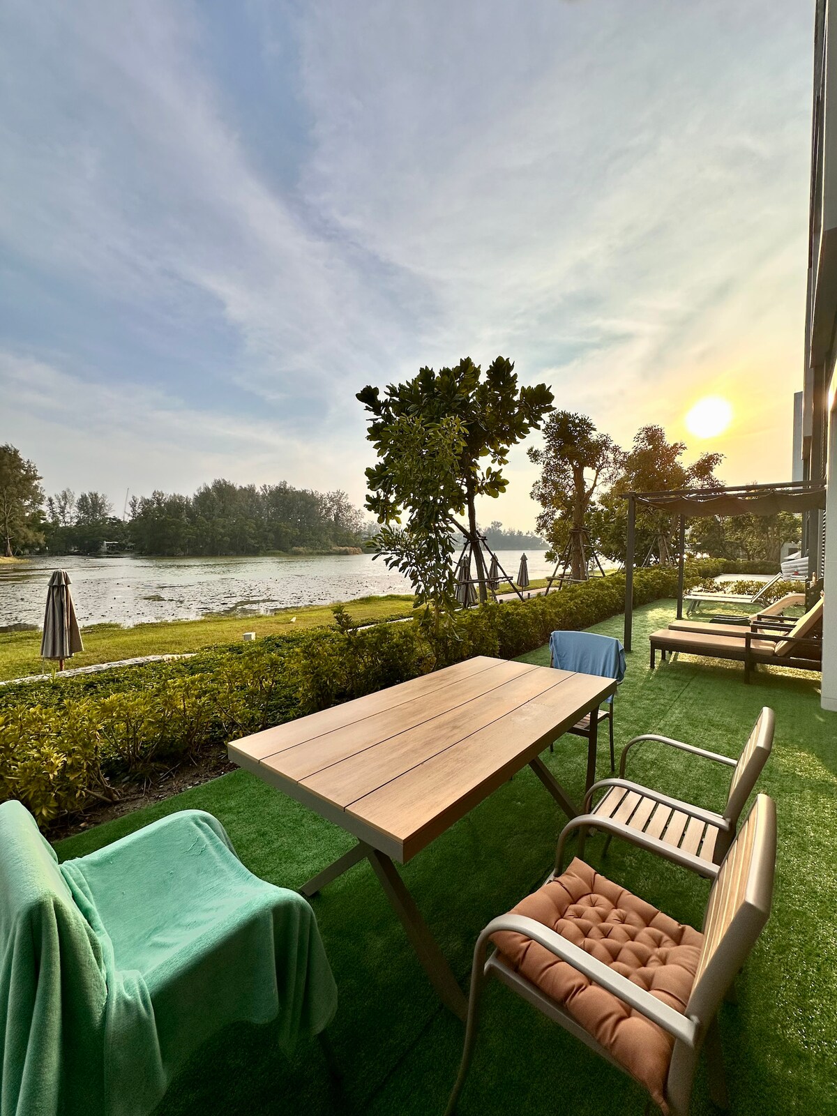 An outdoor patio is set beside a tranquil lake, featuring a wooden table accompanied by chairs. Lush greenery surrounds the area, with lounge chairs positioned for relaxation. The scene is complemented by a soft sunset glow illuminating the horizon, creating a serene ambiance.