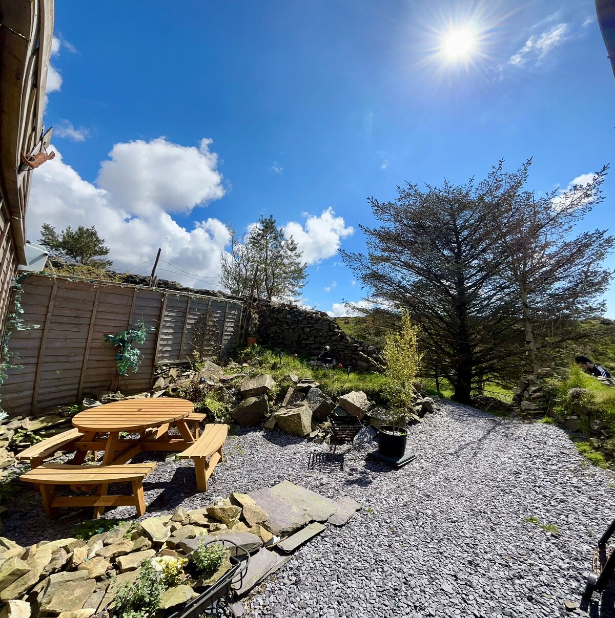 A secluded garden area is presented, featuring a circular table surrounded by benches. Natural stone borders complement the gravel pathway. Shrubs and a tree provide a touch of greenery against a backdrop of blue sky and scattered clouds, inviting outdoor relaxation.