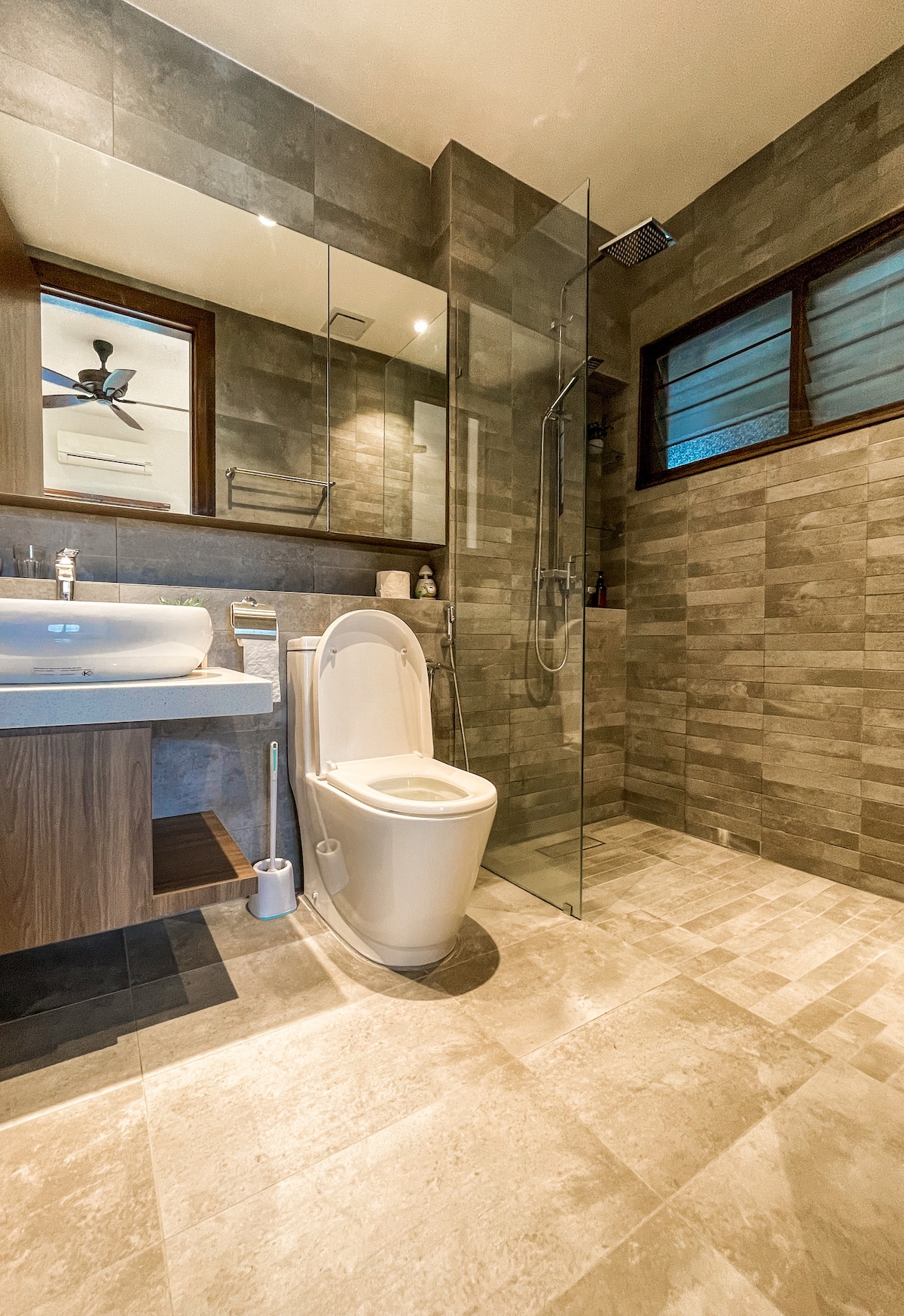 The bathroom features a modern design with a glass shower enclosure and a sleek sink mounted on a wooden vanity. Natural light illuminates the space through two windows, highlighting the textured stone tiles and neutral colour scheme.