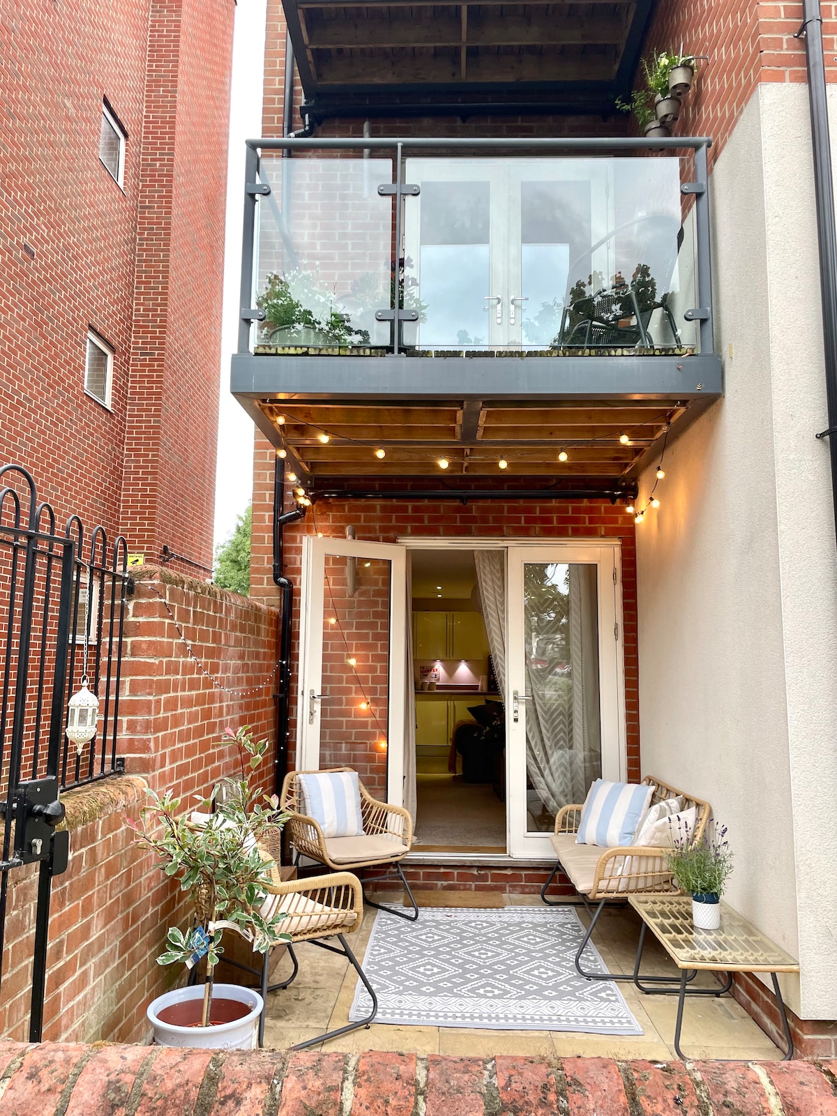 A private patio area features stylish rattan chairs and a small table, positioned on an inviting rug. The space is enhanced by soft lighting and overlooks a communal entrance with greenery along the walls. French doors lead into the open living, kitchen, and dining area.