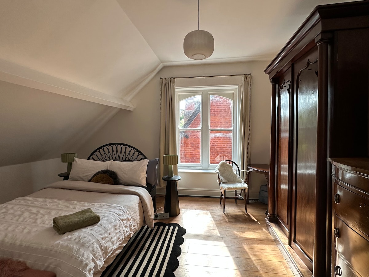 A cozy bedroom is presented with a spacious bed featuring a soft white duvet. A classic wardrobe stands against the wall, and a small armchair is positioned by the window. Natural light illuminates the room, highlighting the wooden flooring and soft furnishings.