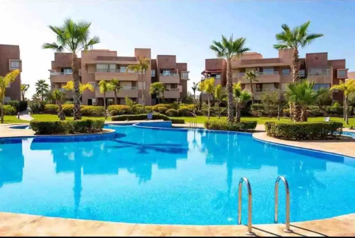 A clear and inviting pool is surrounded by palm trees and neatly trimmed hedges, reflecting sunlight. Buildings with balconies are visible in the background, creating a tranquil and spacious outdoor environment suitable for relaxation.
