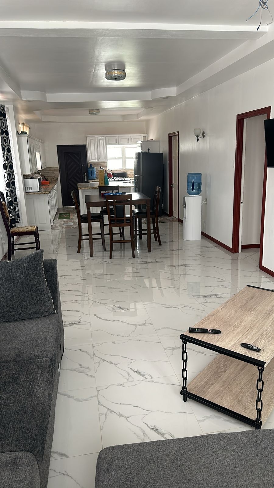 An open-concept living area is highlighted with light-colored walls and modern tile flooring. A dark gray sofa is positioned next to a small wooden coffee table. A dining table with four chairs is visible, along with a kitchen area featuring a refrigerator and cabinetry in the background.