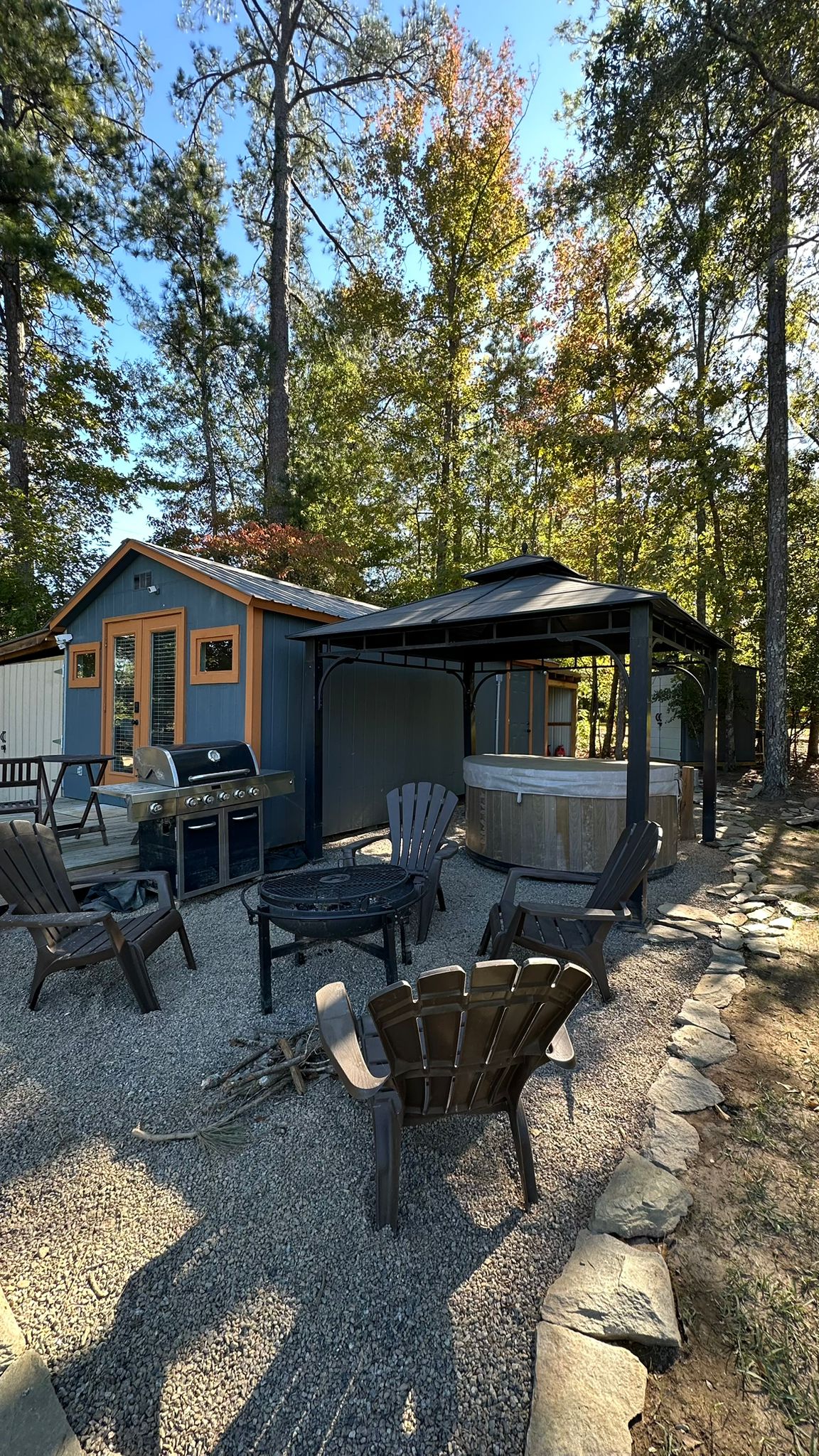 A cozy outdoor space features a gazebo with a hot tub, surrounded by chairs arranged in a circle around a fire pit. A grill is positioned on a nearby platform, with trees providing natural shade in the background.