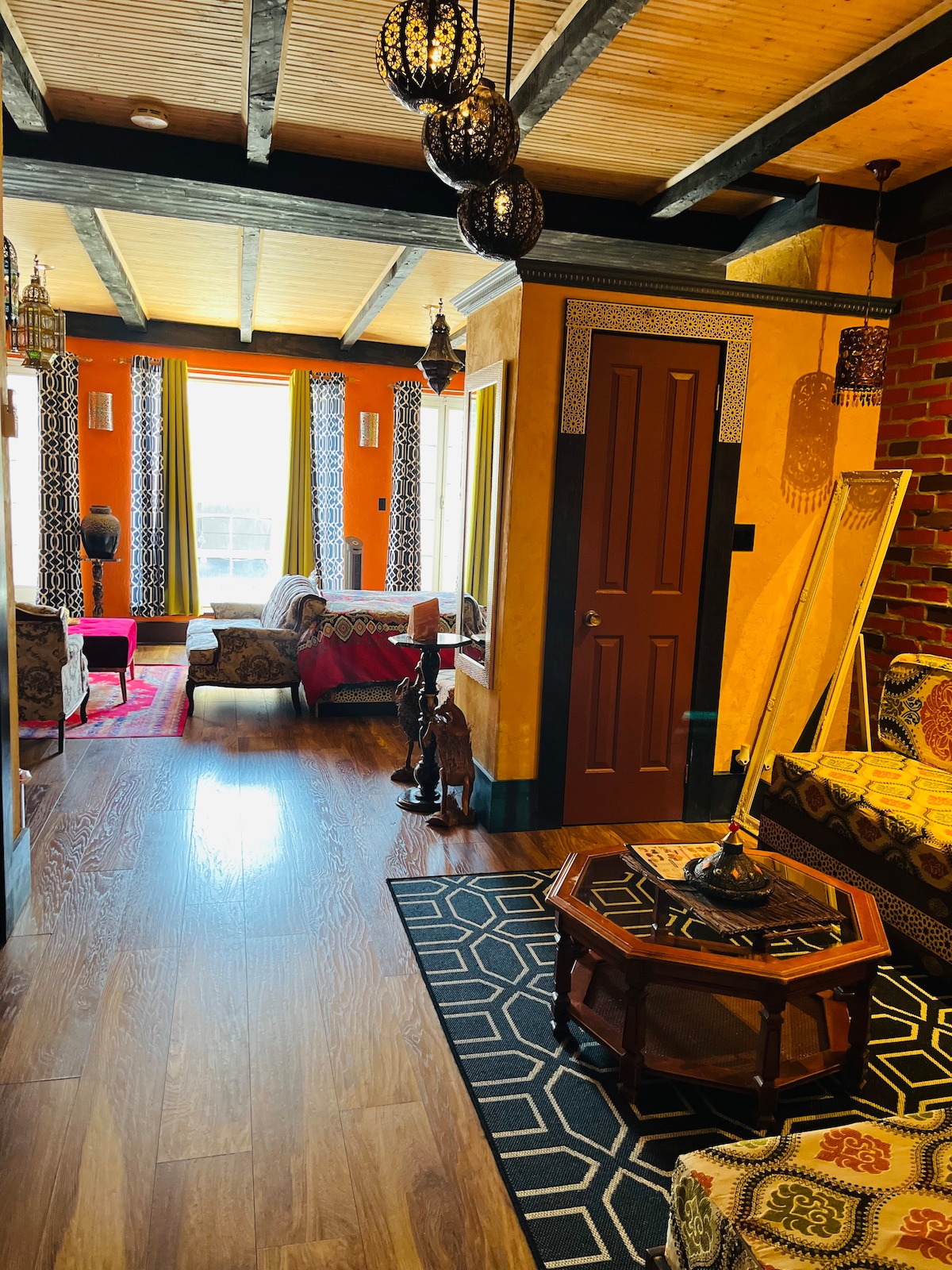 A warm hallway connects various living areas, featuring rich orange walls adorned with patterned curtains. Traditional lanterns hang from the ceiling, casting soft light. Comfortable seating is arranged around a decorative table, and a mirror reflects the inviting space.