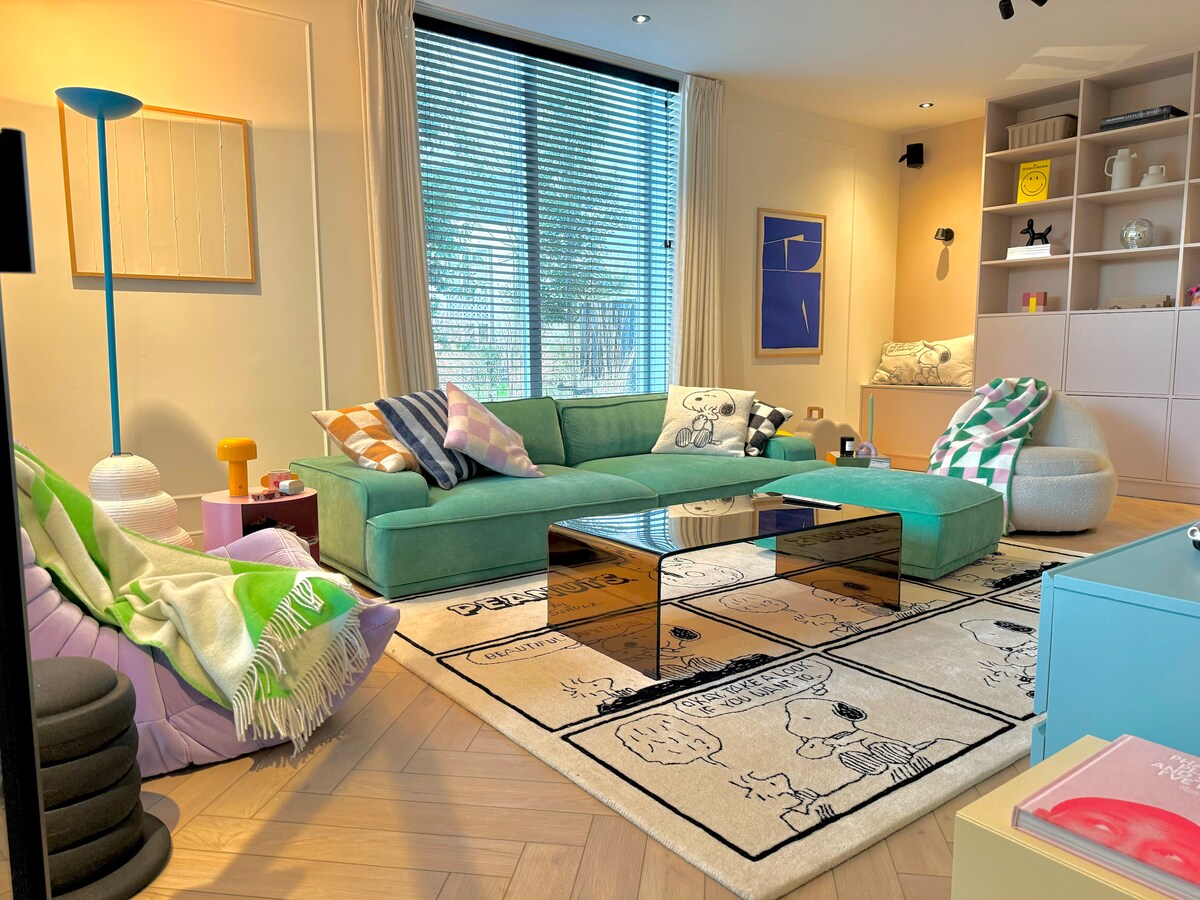 A modern living room is presented, featuring a large green sectional sofa adorned with various cushions. A sleek glass coffee table sits atop a playful area rug. Natural light filters through elegant blinds, highlighting the minimalist decor and vibrant accents in the space.