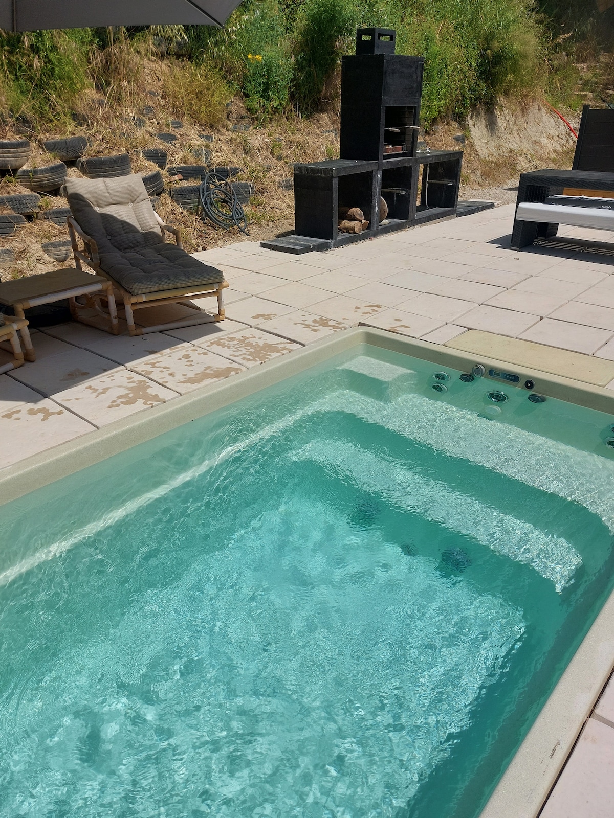 A heated balneo pool is surrounded by a stone patio, enhancing the outdoor space. A pair of lounge chairs offers a spot for relaxation. A barbecue area is visible nearby, complemented by greenery in the background, creating a secluded atmosphere.