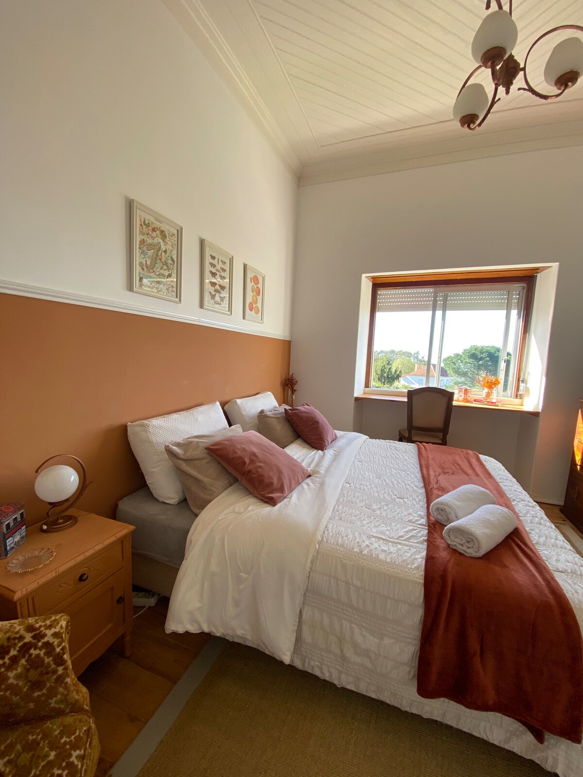 A cozy bedroom features a spacious bed with decorative pillows and soft linens. A bedside table holds a lamp and small decorative items. Natural light floods in from a window, framed by blinds, offering a view of the outdoors. A chair and artwork adorn the walls, enhancing the room's ambiance.
